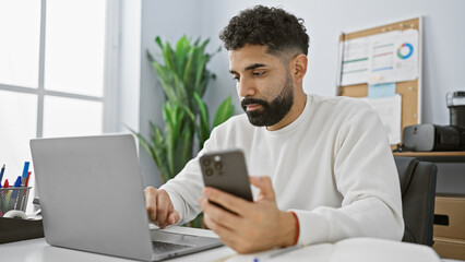 Hispanic man with beard using smartphone and laptop in modern office