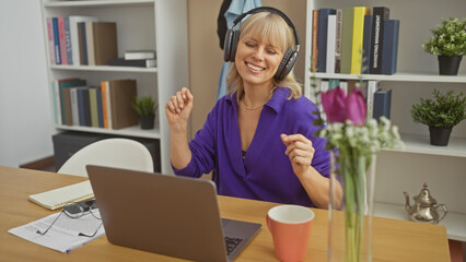 A joyful caucasian woman dances indoors while listening to music on headphones, surrounded by houseplants and a laptop on the table.