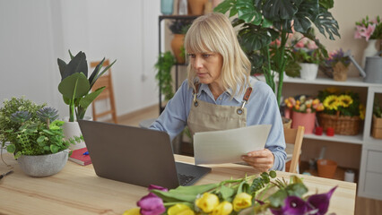Caucasian woman reviewing paperwork in a flower shop surrounded by plants and a laptop