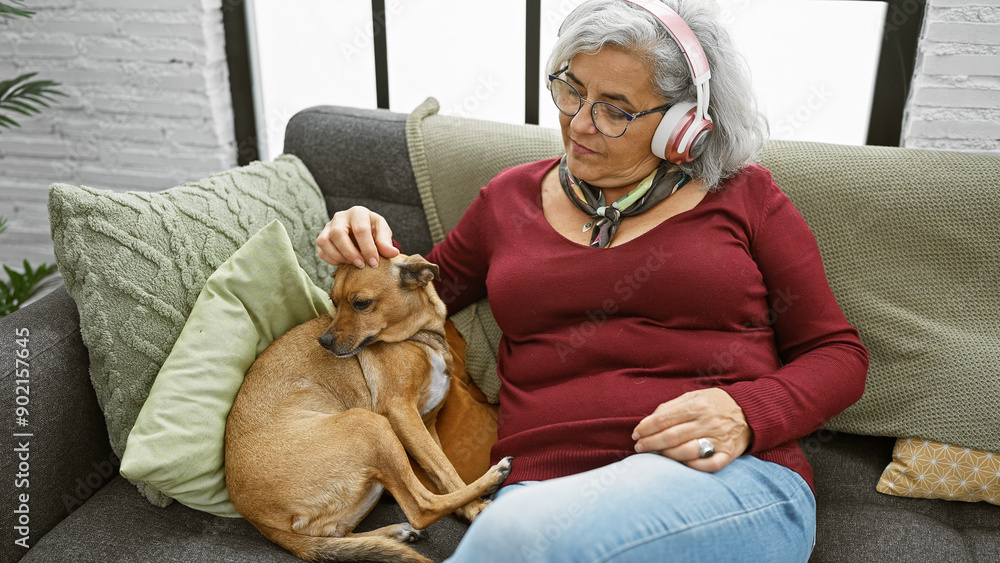 Wall mural A mature woman with grey hair listens to music while petting her dog on a cozy sofa indoors