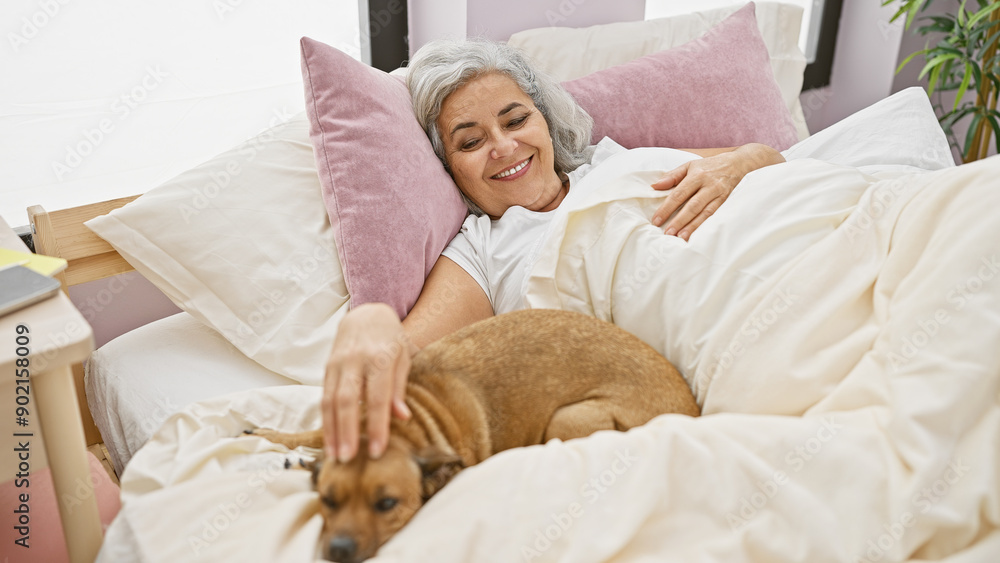 Poster a smiling mature woman lying in bed with a tan dog in a cozy bedroom setting.