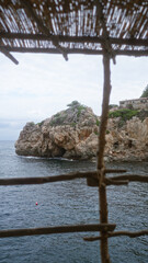 Serene view of the rocky coast of cala deia in mallorca, framed by a rustic bamboo structure with the mediterranean sea in the background.