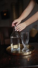 Woman serving water into a crystal glass from a clear bottle on a golden tray in a dimly lit room.