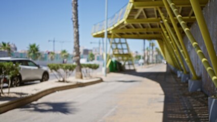 Blurred view of an outdoor urban street with a yellow pedestrian bridge, palm trees, and a parked car on a sunny day, capturing an abstract city scene with defocused elements.