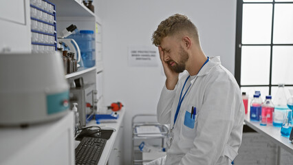 Naklejka premium Stressed young man in lab coat standing in a laboratory with his hand on his forehead, looking overwhelmed.