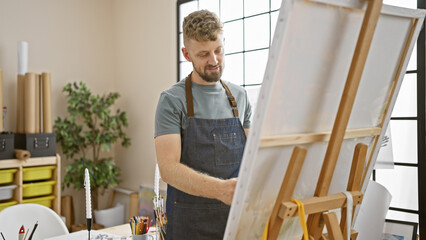 A handsome young caucasian man with a beard and blue eyes wearing an apron stands before an easel in a sunny art studio.