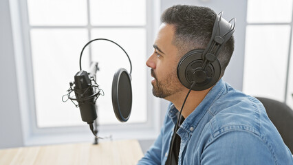 Handsome hispanic man wearing headphones speaking into a microphone in a bright, modern radio...