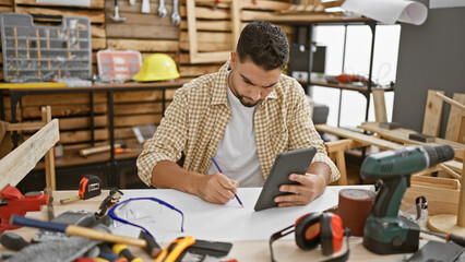 A bearded man indoors uses a tablet while planning in a cluttered woodworking workshop.