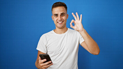 Handsome young man with smartphone making okay gesture against blue wall