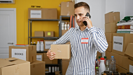 Young hispanic man on phone in warehouse, holding box, with labeled shelves and donations background.