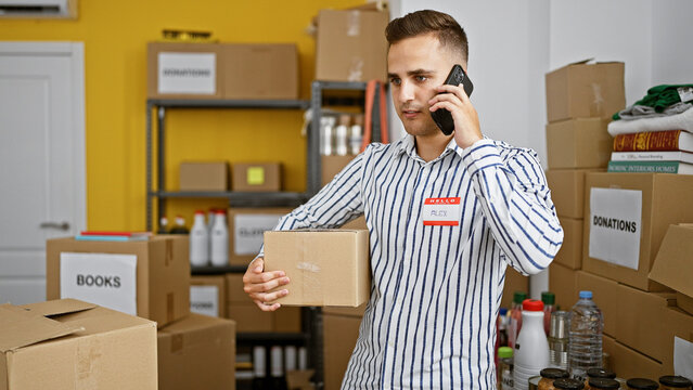 Young hispanic man on phone in warehouse, holding box, with labeled shelves and donations background.