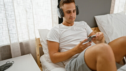 Fototapeta premium A young man relaxing in bed with headphones and a smartphone in a cozy bedroom setting