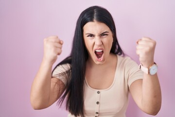 Young hispanic woman standing over pink background angry and mad raising fists frustrated and furious while shouting with anger. rage and aggressive concept.