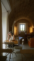 Young hispanic woman sitting indoors at a cozy rustic restaurant or hotel cafeteria, enjoying a peaceful moment by the window with a cup of coffee.