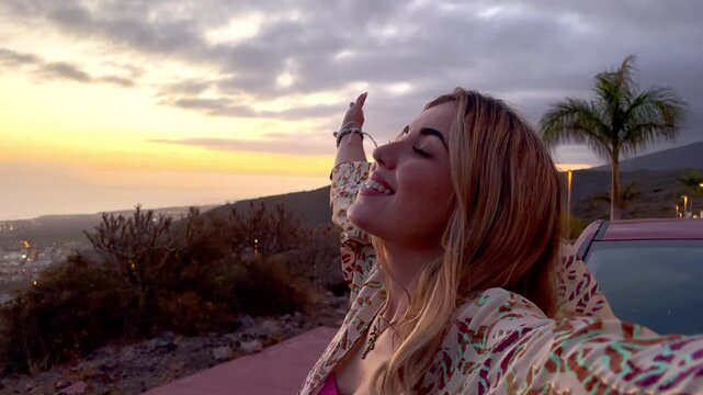 Young beautiful woman enjoying summer and having fun sitting on the hood of the car opening arms feeling good and free. Freedom concept and happy lifestyle at sunset
