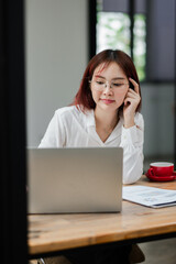 Focused young woman in glasses working on a laptop at a modern office desk with a coffee cup and documents.