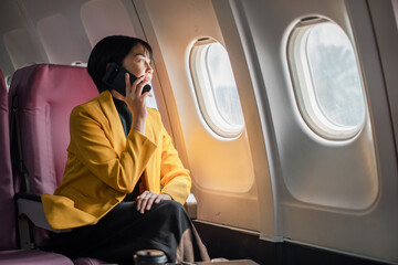 Professional woman in yellow blazer using smartphone on airplane, gazing out window. Business travel concept.