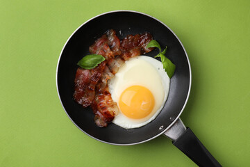 Fried egg, bacon and basil in frying pan on green background, top view