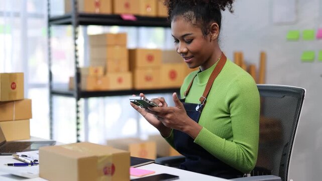 Small business owner checks her smartphone while efficiently packing products in an organized workspace, showcasing a modern and effective business operation.