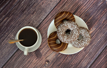 A cup of hot black coffee and a plate of fresh donuts with chocolate caramel glaze on a dark wooden table.