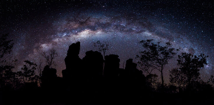 Milky Way galaxy arching over a silhouette of rocky terrain and sparse trees.