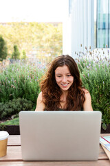 Front view of happy young redhead female university student using laptop outdoors sitting on table studying. Vertical