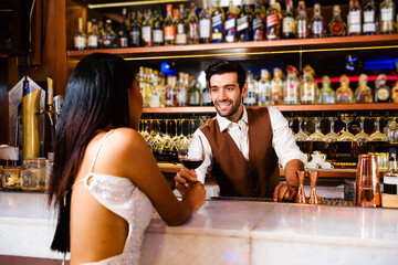 Caucasian professional bartender or mixologist making a cocktail for women at a bar. An attractive barman served a glass of wine with a smile to a woman. Bartender service at the night club restaurant