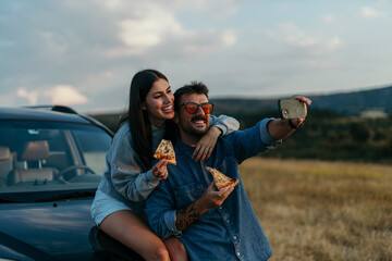 Loving couple taking a selfie outdoors, leaning on the hood of their car in the evening, eating fast food