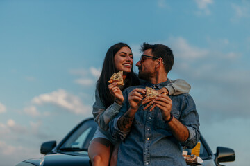 Young heterosexual couple in nature chilling next to the car and having fun eating junkie food