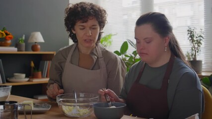 Girl with Down syndrome wearing apron sitting at table, adding sugar to butter and mashing ingredients together in bowl under guidance of female teacher during cooking class