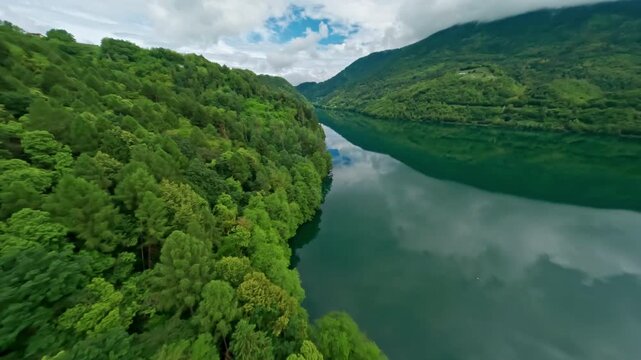Lush green forest surrounds the serene Lago di Levico, reflecting the cloudy sky, aerial view