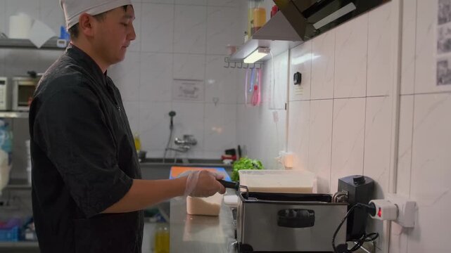 Side view of asian chef holding deep fryer frying food in boiling oil while standing in commercial restaurant kitchen.