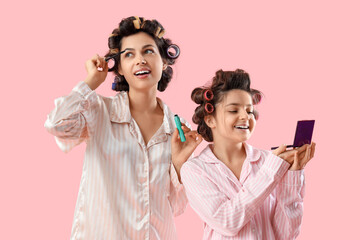 Little girl and her mother with hair curlers in pajamas doing makeup on pink background