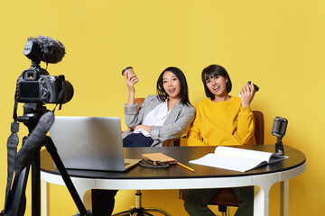 Young women with coffee recording podcast at table on yellow background