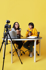 Young women with notebook and coffee recording podcast at table on yellow background