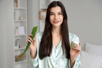 Beautiful young happy woman with plant leaf and jar of aloe vera gel in bedroom