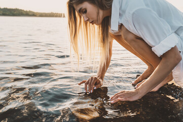 Beautiful blonde woman touching water in river on sunset in golden sun beams. scoop up water in palms simple natural fairytale photography. Noise blurred image