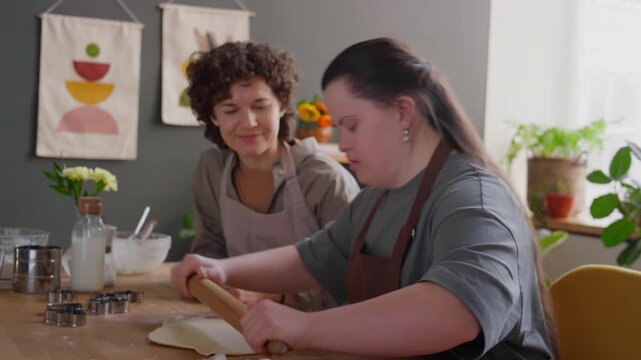 Girl with Down syndrome learning hot to roll pastry dough with assistance of female tutor during cooking lesson