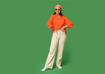Cool African-American woman wearing hat and sunglasses on green background