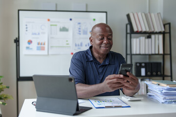 Retired African American male businessman sitting talking on mobile phone in office using laptop computer online. Contacting business customers, communication, various document work.