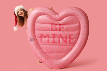 Happy young woman in swimsuit and Santa hat with inflatable mattress on pink background. Christmas...
