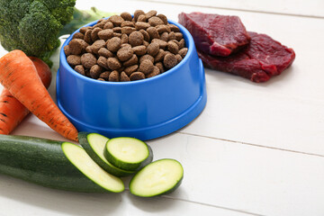 Bowl with healthy dry pet food near vegetables and meat on white wooden background