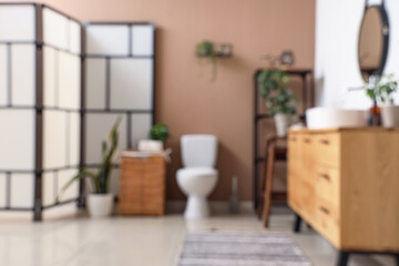Modern toilet bowl, folding screen and chest of drawers near brown wall in restroom. Blurred view