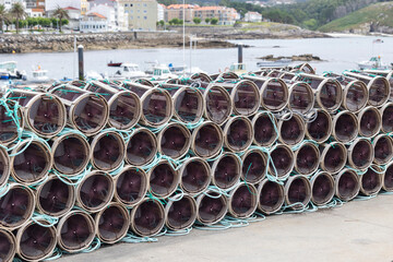 A large stack of cylindrical fishing traps is neatly arranged beside a waterfront dock, ready for...
