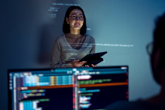 asian woman developers collaborating on a coding project, with one member presentation at code projected on the wall while the other works on multiple computer screens