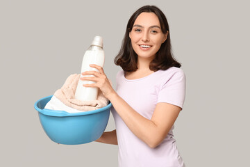 Young woman with laundry basket and bottle of detergent on grey background