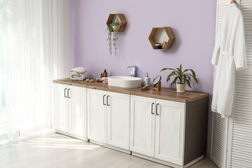 Folding screen, chest of drawers with sink and bath supplies near lilac wall in interior of bathroom