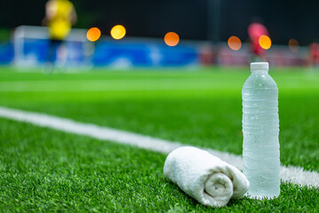 Close-up of a water bottle and towel on a soccer field, with blurred players in the background during night practice. © chartphoto