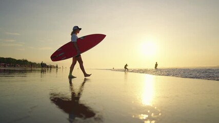 Athletic young woman with surfboard in hands walking on sandy beach to ocean. Merry summer vacation, slow motion shot, female silhouette against bright sunshine reflecting in water, early morning - Powered by Adobe