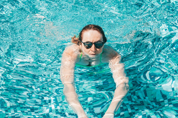 Active mature caucasian woman swimming in outdoor swimming pool under the sunlight wearing sunglasses. Vacation and healthy lifestyle concept